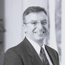 Black and white photo of person wearing glasses and suit and tie.