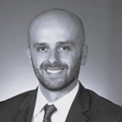 Photo shows black and white headshot person in suit and tie in front of a dark background.